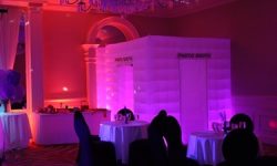 Our enclosed photobooth lights up eight different colors and is 8'X8'. A glowing inflatable photo booth is set up in a dimly lit event room with tables and chairs nearby.