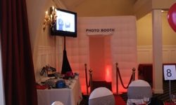 Here we had our VIP photobooth package which includes our enclosed booth, one- (1) TV for a slideshow of the fun inside and our red carpet with gold posts and red ropes to make you feel like a VIP. White inflatable photo booth with red lighting, rope barriers, and a table of props in an event room.
