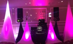 Here is our Walker Package. This includes our 18 wireless up-lights with color of your choice. Transform any room into a magical setting. The light can also change colors or dance to the beat of the music for a very cool effect. DJ booth setup with speakers, lights, and pink uplighting in an indoor event space with patterned carpet.