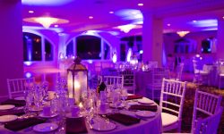 Elegant banquet hall with round tables, white chairs, and purple lighting; lantern centerpiece on foreground table.