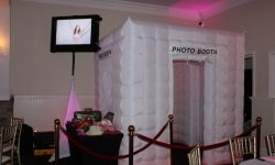 An inflatable white photo booth is set up in an event space, with props on a table and a monitor displayed above.